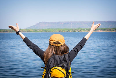 Rear view of backpack woman with arms raised standing by lake against clear sky