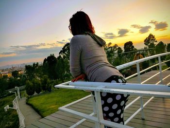 Rear view of woman sitting on railing against sky during sunset