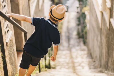 Rear view of boy wearing hat standing on staircase by railing outdoors