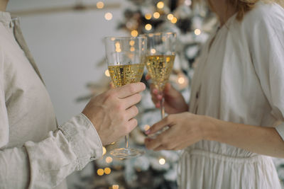 Midsection of couple holding champagne flutes while standing by christmas tree at home