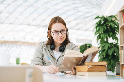 Young teenager girl college student in glasses doing homework with books at green modern library 