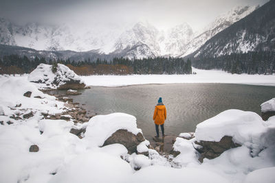 Rear view of woman standing at lakeshore against snowcapped mountains