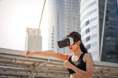 Young woman using virtual reality simulator punching against building in city