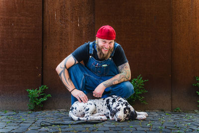 Happy bearded man with tattoos wearing denim overall and cap and sitting on cobblestone pavement with cute spotted great dane puppy