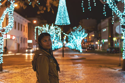 Thoughtful young woman standing on road against illuminated christmas decoration in city at night