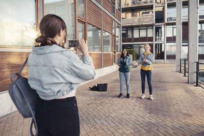 Teenage girl photographing female friends while standing on footpath against building