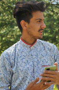 Closeup of a indian young guy standing outdoor, holding mobile phone in his hands with looking away