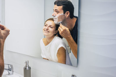 Woman looking at man shaving while standing in front of mirror