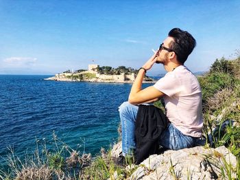 Young man sitting on rock by sea against sky