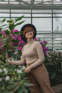 A beautiful plus size girl in a hat and glasses smiles among the green plants of the greenhouse. 