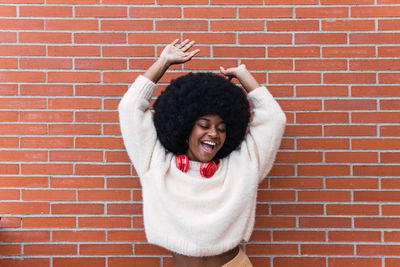 Young woman with arms raised standing against brick wall