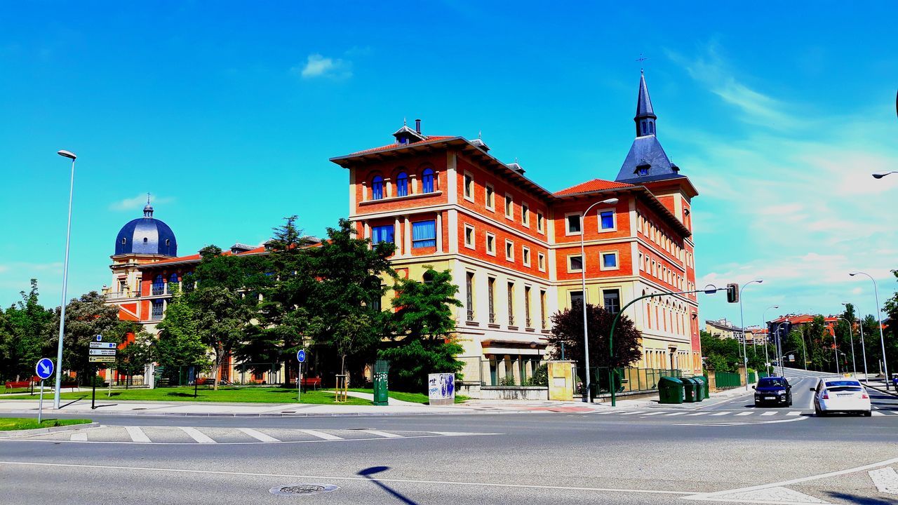 architecture, building exterior, city, built structure, street, transportation, sky, road, mode of transportation, building, car, nature, motor vehicle, tree, incidental people, land vehicle, plant, day, blue, city street, outdoors, spire