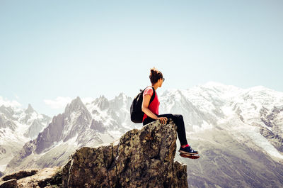 Young woman looking away while sitting on rock against sky