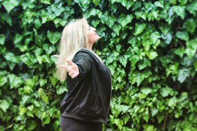 Side view of woman with arms outstretched standing against plants in park