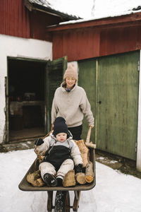 Smiling mother carrying daughter sitting on firewood in wheelbarrow during winter