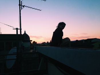 Silhouette woman sitting on retaining wall against sky during sunset