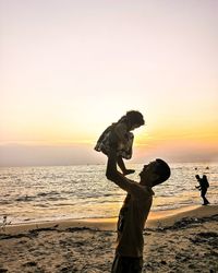 Side view of man holding aloft baby girl at beach during sunset
