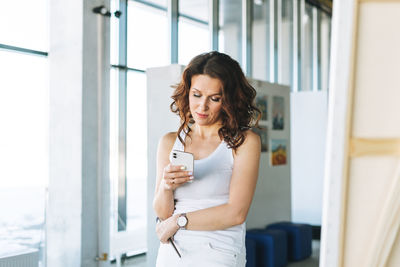 Thinking woman artist with paint brush using mobile in front of easel in art gallery