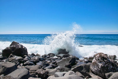 Waves splashing on rocks at shore against clear sky