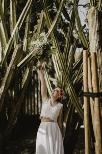 Gleeful young woman in white top and skirt smiling and raising arm with bridal bouquet while standing near tall cacti on wedding day in garden