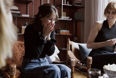 Surprised woman sitting on chair next to female friend in apartment