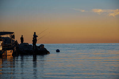 People fishing in sea against sky during sunset