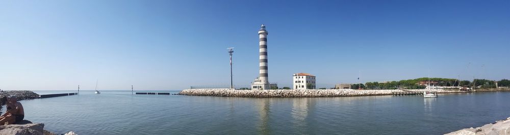 Lighthouse by sea against clear blue sky