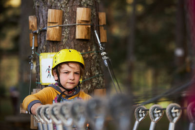 Boy in yellow helmet standing on suspension bridge near tree trunk on summer weekend day in adventure park in forest