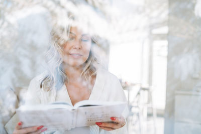 Young woman with blonde curly hair in white cardigan reding book near window with winter landscape