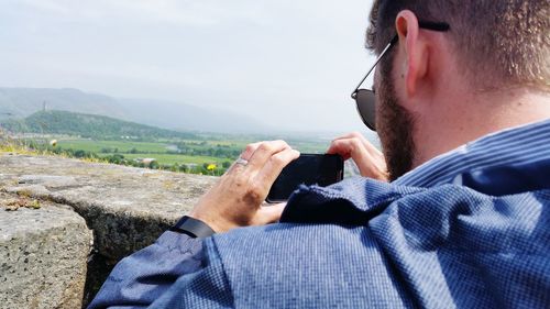 Rear view of man photographing landscape through mobile phone against sky