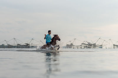 Man riding horse in lake against sky during sunset