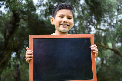 Portrait of young woman holding blackboard against trees