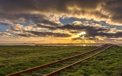 Railroad tracks on field against cloudy sky during sunset