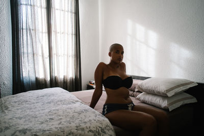 Calm curvy black female sitting on soft bed in modern bedroom and looking at camera while resting at home