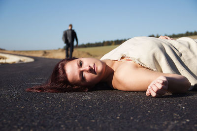 Portrait of young woman lying down on road against clear sky