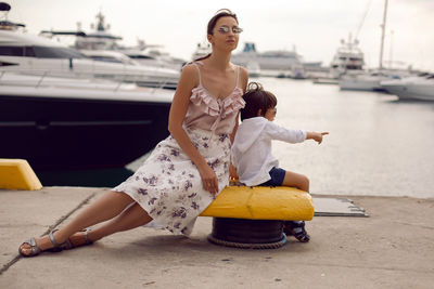 Mother in a dress with her son sitting on the pier with yachts on the sea in the summer