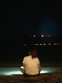 Rear view of woman sitting on beach against sky at night