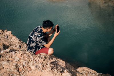 High angle view of young man photographing with mobile phone while crouching on rock by lake