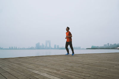 Mature man running on pier over lake against sky in city