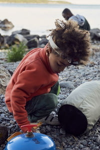 Full length of boy crouching by bag on rocks at beach