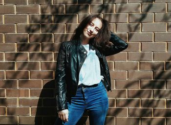 Portrait of smiling woman with hand in hair standing against brick wall