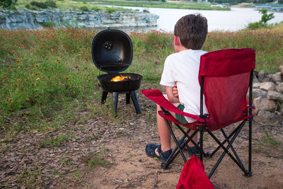 Rear view of boy sitting on chair with burning camping stove