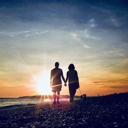 Rear view of couple holding hands at beach against sky during sunset