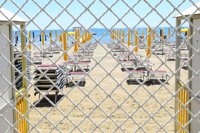 Deck chairs with parasols arranged at sandy beach seen through fence during sunny day