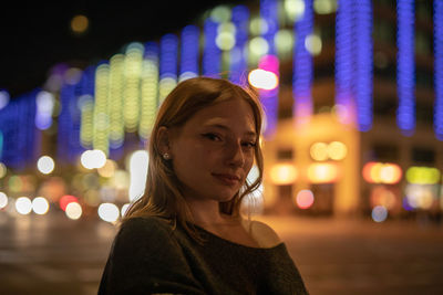 Portrait of young woman standing against illuminated city at night