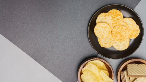 Flat lay of snack, crispy potato chip on black plate and wafer in wooden bowl over grey background.