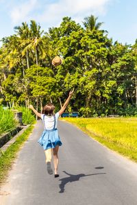 Rear view of woman catching hat while running on road against trees