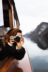 Unrecognizable female photographer in warm jacket shooting with photo camera while travelling by boat on lake with rocky shore in autumn day