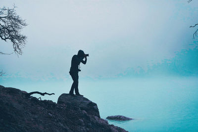 Silhouette young man photographing while standing on rock nu sea during foggy weather