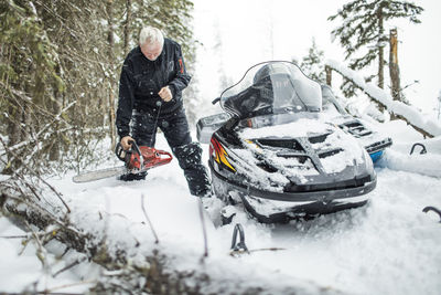 Retired man using chainsaw to clear trails while snowmobiling.